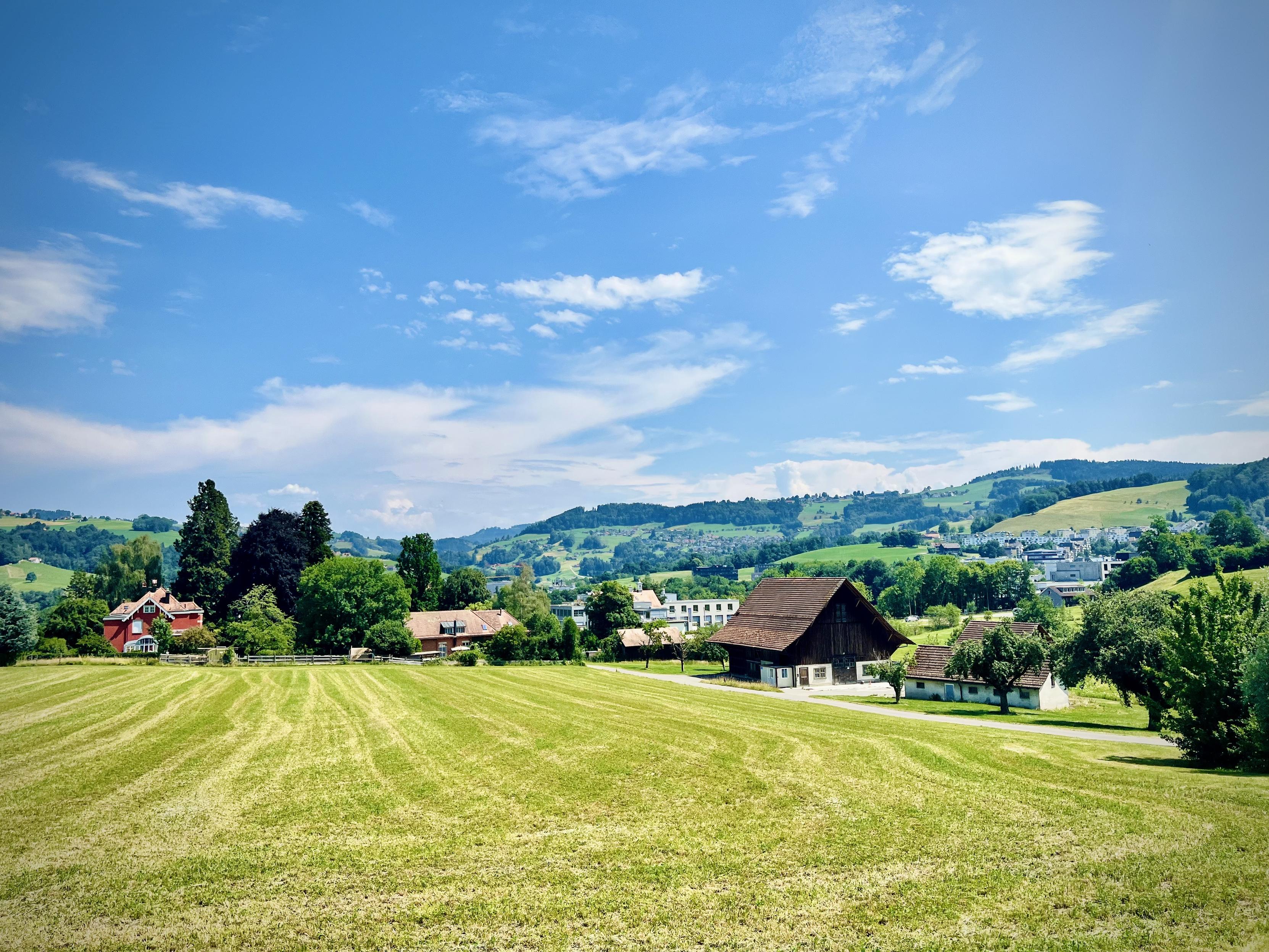 A scenic rural landscape under a bright blue sky with light, scattered clouds. In the foreground, a large, mowed grassy meadow stretches toward a few traditional farmhouses and scattered buildings. Rolling green hills filled with trees and more distant houses rise in the background, creating a peaceful, countryside atmosphere.