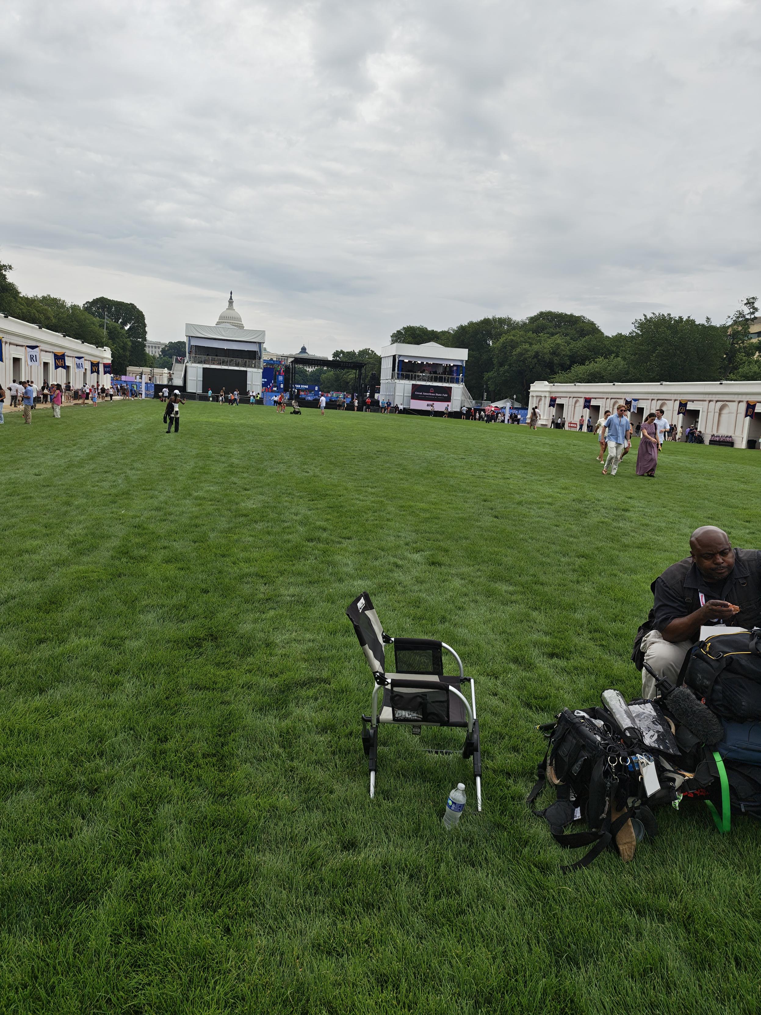 A professional photographer sitting, semi slumped over his pile of gear, looking bored. In the background there are a couple of dozen people near the temporary booths set up for the event.  The expanse of grass behind him looks like empty pasture.