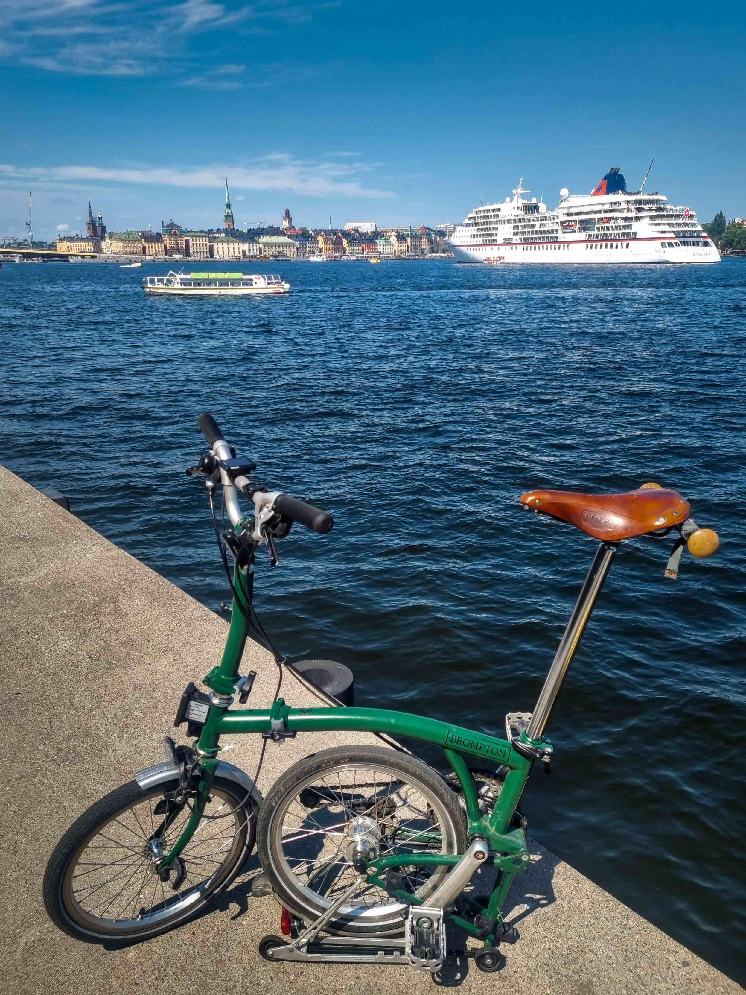 A folding bike next to the harbour of Stockholm, Sweden. A huge tourist boat behind. In the far, the Old Town of Stockholm.