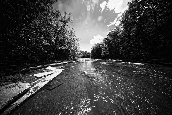 High contrast monochrome image of shallow creek running over shaly flat rocks with walls of dark trees on both sides and dramatically lit clouds