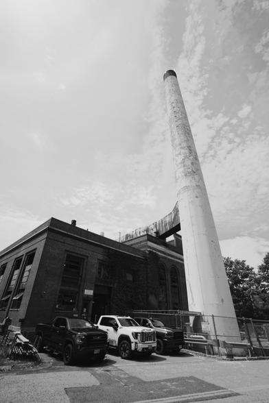 White smokestack rises next to squat dark brick building with pickup trucks parked by it under a huge sky of (I think) moderate altitude clouds