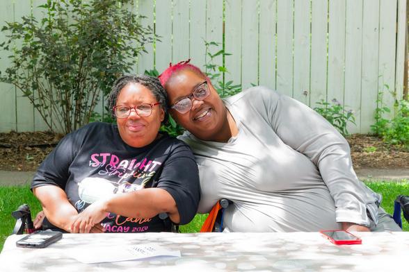 Two dark skinned women lean in together with their phones in front of them on a white table with a white fence and green vegetation behind them