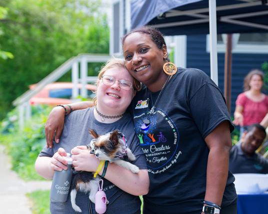 On the left a shorter white woman holds a little dog,  on the right a taller black lady has her arm around the first and a big earring that says "Black Beauty".  Bothj are smiling and wearing black shirts.
