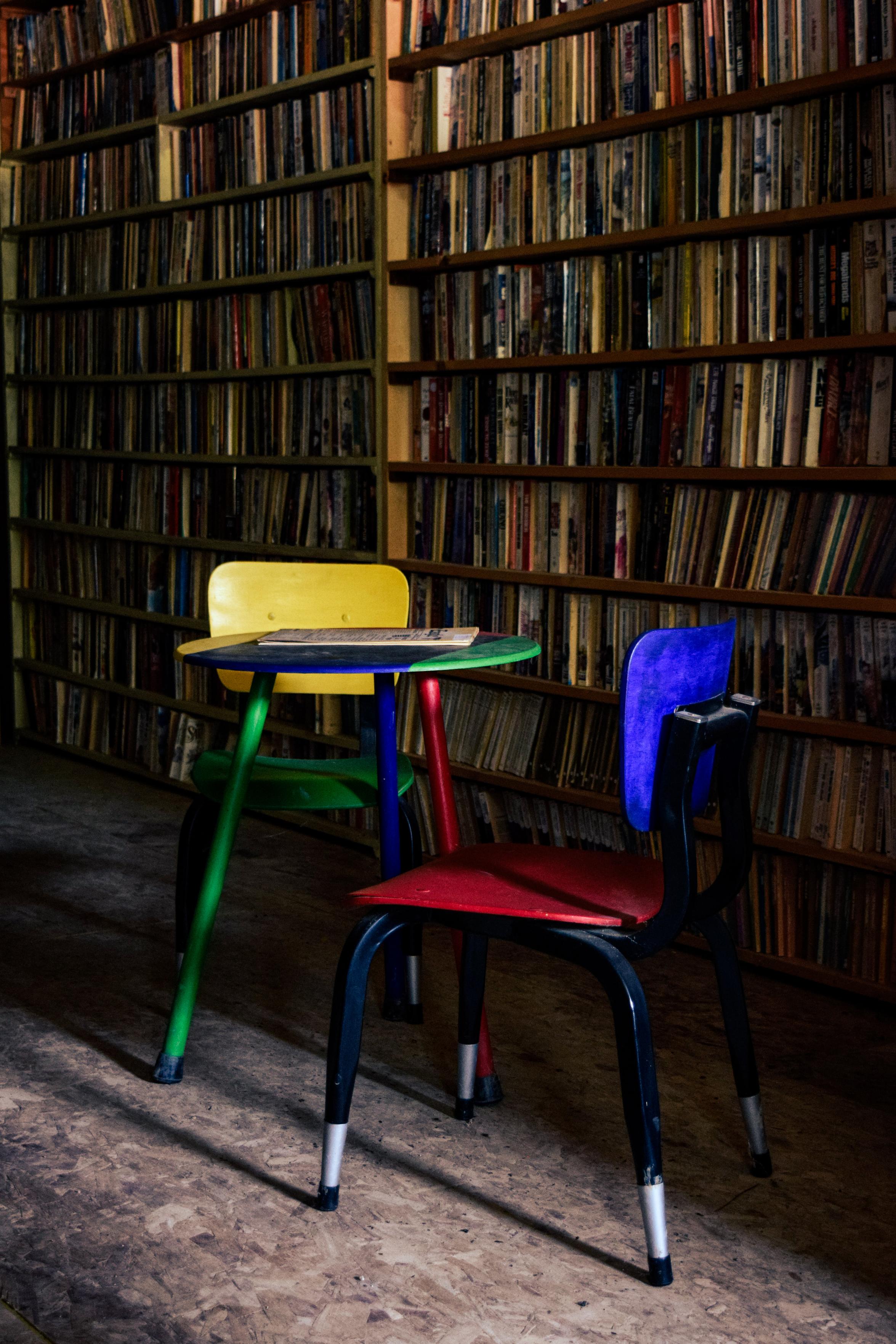 Colorful children's table and chairs set against shelves filled with rows of books. The scene is warmly lit, creating a cozy library atmosphere.