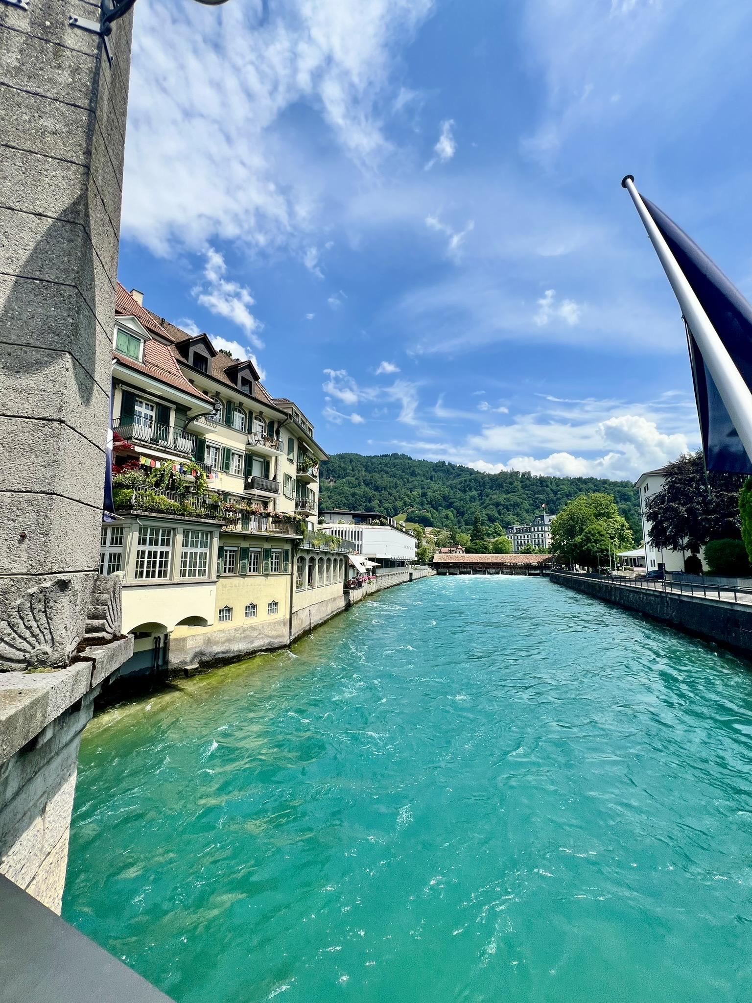 A vibrant turquoise river flows through a city canal, flanked by multi-story buildings with balconies and green shutters on the left and a stone embankment on the right. In the distance, a covered wooden bridge spans the river, backed by a densely forested green hill under a bright blue sky filled with white clouds.