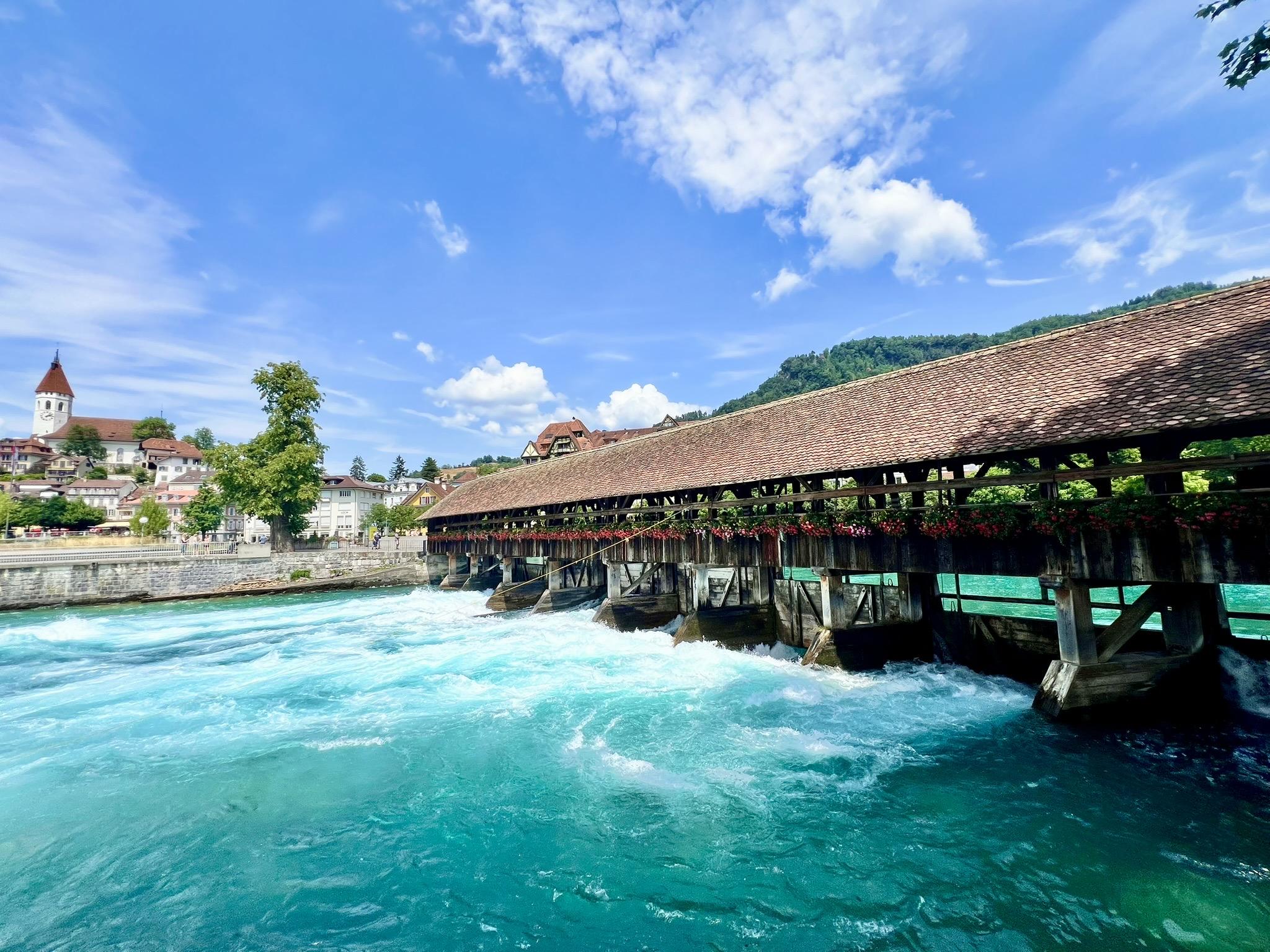 A historic covered wooden bridge decorated with boxes of red flowers spans a rushing, churning turquoise river. On the left bank, a town rises on a hill, featuring a prominent white church tower with a red-pointed roof, while a forested green hill rises on the right under a bright blue sky with scattered white clouds.