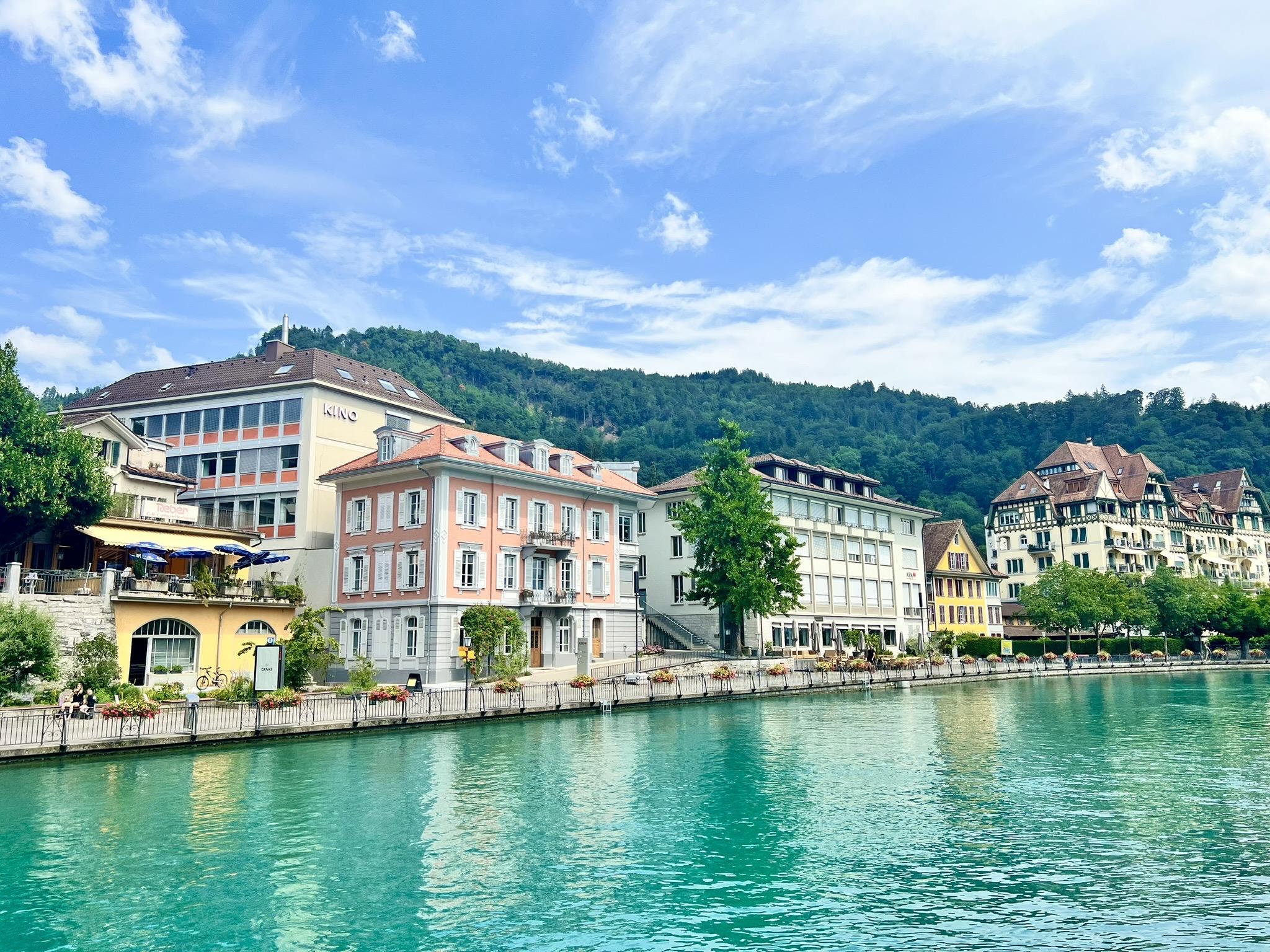 A row of colorful, multi-story buildings, including a prominent salmon-pink structure with white shutters and a traditional timber-framed house on the right, lines a waterfront pedestrian promenade decorated with flower boxes. In the foreground, a vibrant turquoise river flows past, while a steep, densely forested hill rises in the background under a blue sky with light white clouds.