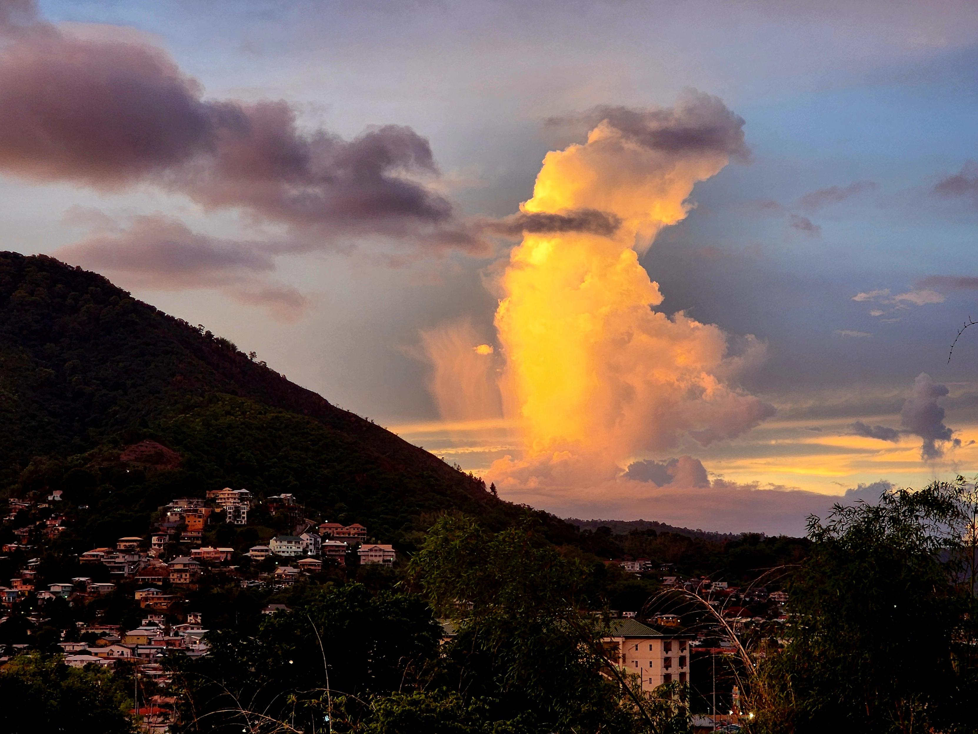 A sunset illuminates a towering cumulonimbus cloud in brilliant gold and orange beyond a forested hillside overlooking a small town. Dark gray clouds drift across the glowing cloud's upper section, creating striking contrast, while the foreground sits in deep shadow with scattered houses nestled along the hillside. The low evening light transforms the storm cloud into a luminous column rising high into the sky against soft blues, purples, and warm amber hues.