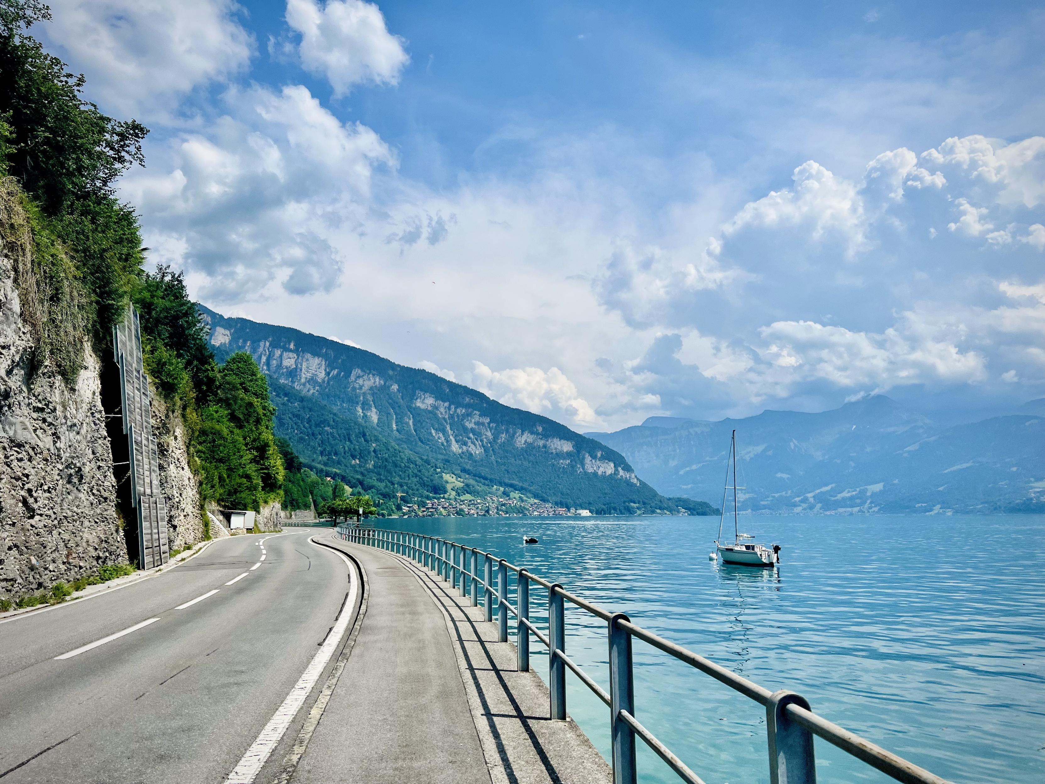 A paved two-lane road and adjacent bicycle path curve along a turquoise lake, bordered by a steep rocky cliff on the left and a metal railing on the right. A single small white sailboat floats in the calm water, with steep, forested mountains rising in the background beneath a bright blue sky filled with dramatic white clouds.