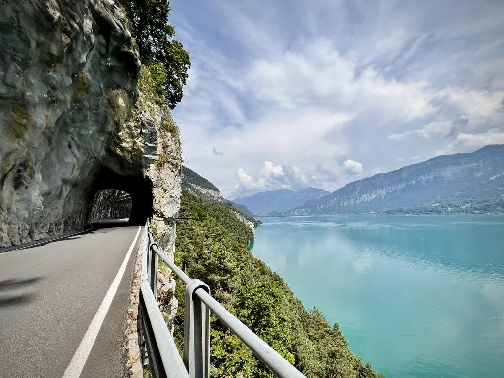 A paved road on the left leads into a dark tunnel carved through a sheer, rugged rock cliff, bordered by a silver metal safety railing. To the right, the cliff drops off into a dense forest that meets a calm, vast turquoise lake, with majestic mountains rising in the distance under a light, cloudy sky.