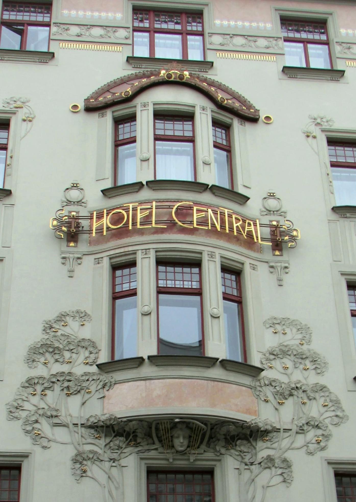 Low light winter's day photo of some of the front exterior of The Hotel Central in Prague. It shows 2 curved bay windows, one above the hotel sign and one below. The hotel frontage is painted pale green and pink with gold. Below the lower window there is a carved female face and graceful, stylized trees decorate the wall on either side of the window. The curved sign between the windows says "HOTEL CENTRAL" in golden capital letters.