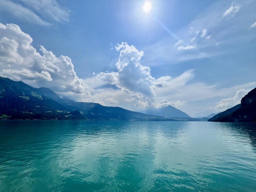 A wide expanse of calm turquoise water is framed by steep, dark green mountain shores on both sides. The bright sun shines high in a blue sky, casting a soft glare over towering, dramatic white cloud formations that rise above the distant mountain peaks.