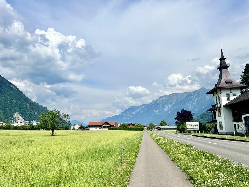 A straight asphalt pathway runs parallel to a paved road, bordered by an open, light green field with a solitary tree on the left. On the right, next to the road, stands a white, multi-story building with a prominent, dark pointed steeple-like roof, all set against a backdrop of majestic mountains and dramatic clouds.