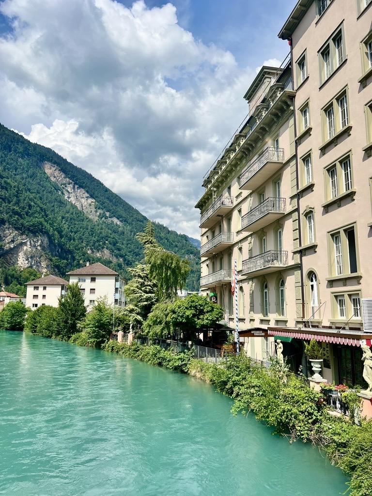 A vibrant turquoise river flows past a grand, multi-story building with classic iron-railed balconies on the right bank. Lush green vegetation lines the water's edge, leading to a steep, forested mountain rising in the background under a partly cloudy sky.