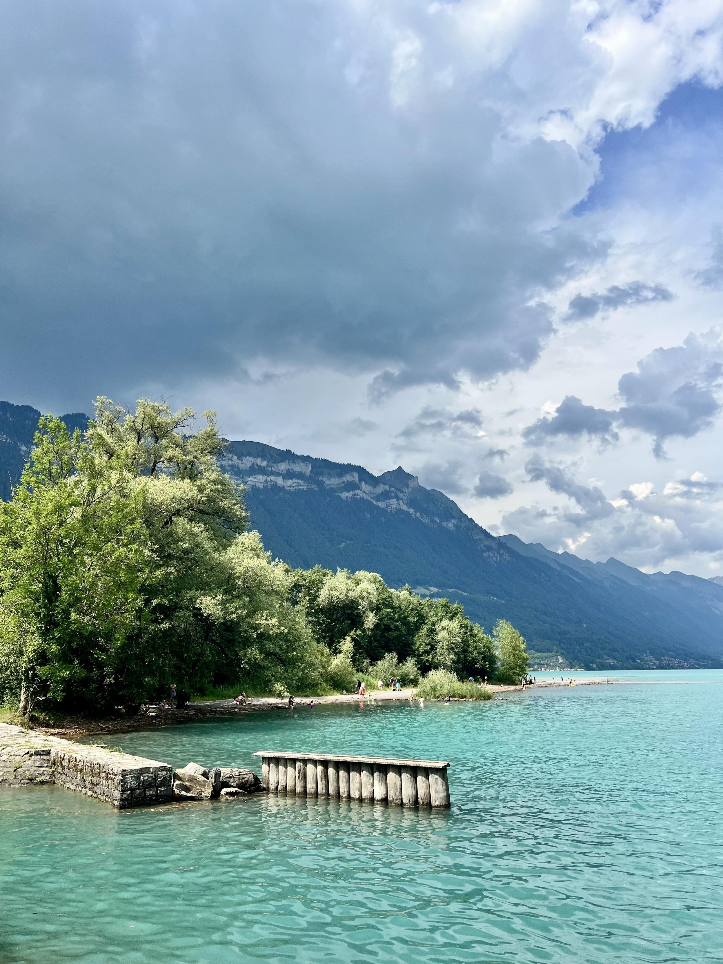 A small stone and wooden jetty extends into a vibrant turquoise lake, with a tree-lined shoreline and a small beach where people are gathered on the left. In the background, dramatic, dark storm clouds hang over a steep mountain range.