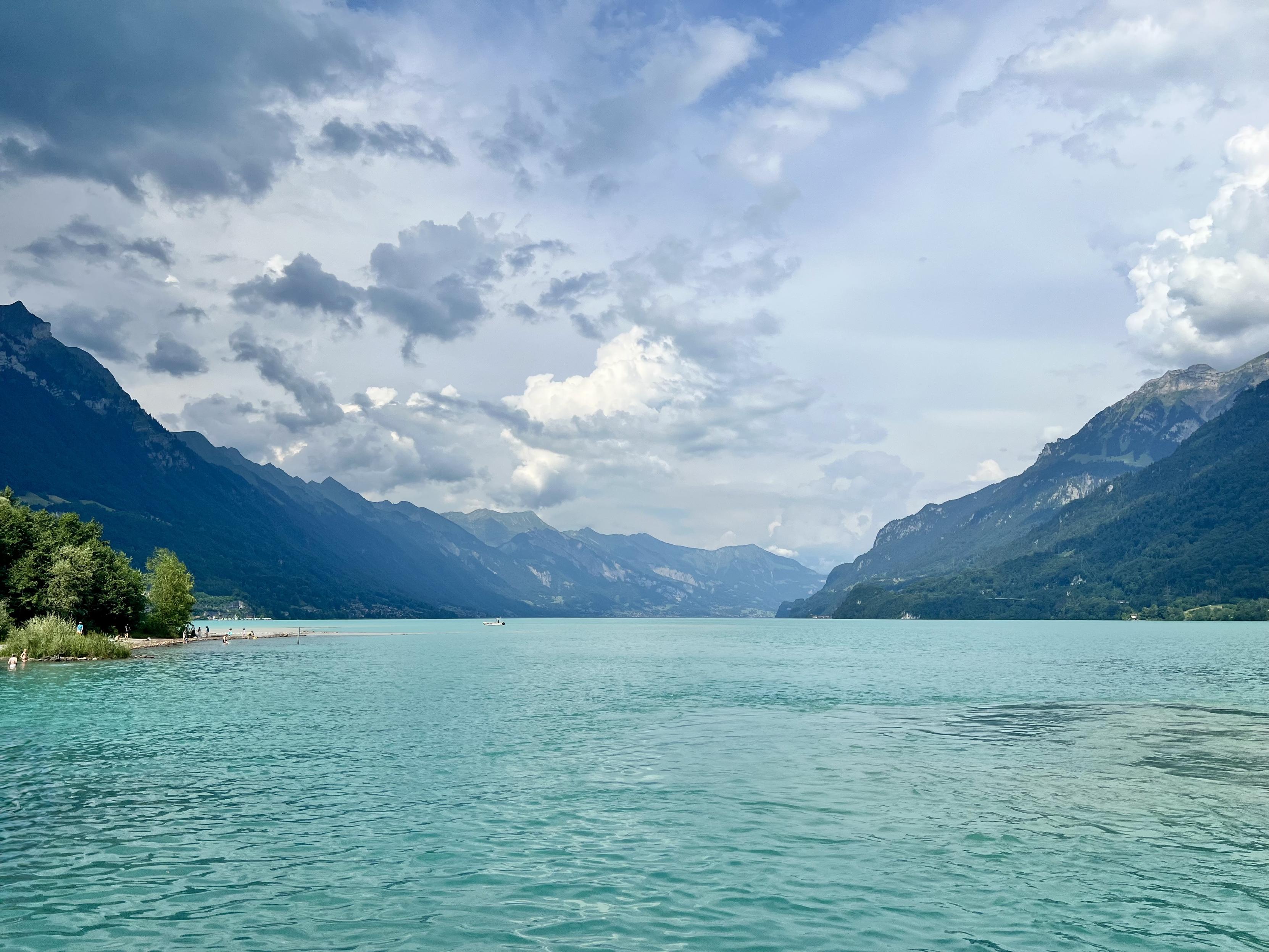 A wide perspective of a vibrant turquoise lake stretching between two steep, forested mountain ranges under a partly cloudy sky, with a small tree-lined shore visible on the far left.