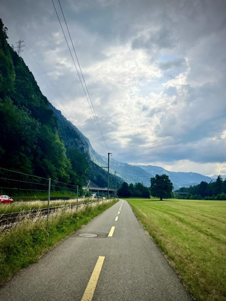 An asphalt bicycle path with a dashed yellow center line runs parallel to a railway track and a steep, forested mountain slope on the left, with a flat green field extending to the right under a cloudy sky.