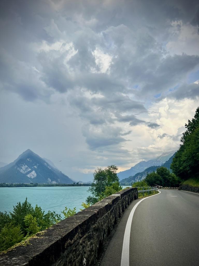 A paved road curves along a low stone retaining wall overlooking a bright turquoise lake, with a prominent, pointed mountain in the distance under a cloudy sky.