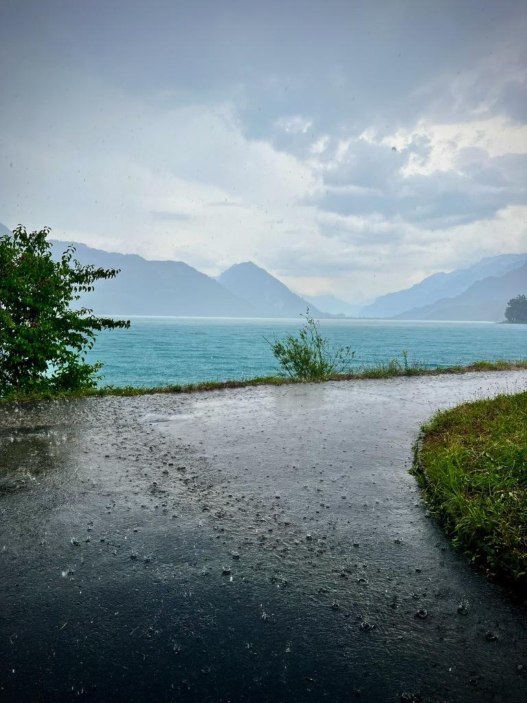 Heavy rain falls on a wet asphalt path, creating ripples on its reflective surface, overlooking a turquoise lake with misty, distant mountains under a grey sky.