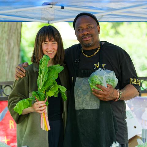 White woman with shoulder-length hair in a bob haircut and a long green jacket and dark shirt on the left holds a bunch of greens with colorful stems while a black man on the right has a hand around her shoulder, a black shirty, a gold chain and holds a bag of lettuce or arugula or somethin underneath a tenth with bright blurry foliage in the far background