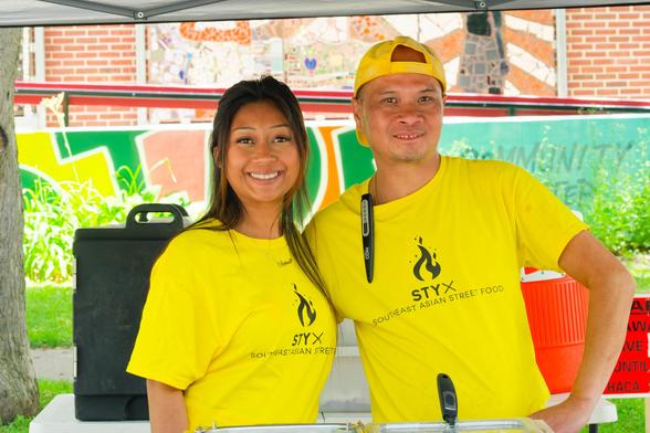 Smiling asian woman (left) and man (right) wear bright yellow shirts that read STYX: SOUTHEAST ASIAN STREET FOOD and the man has a yellow backwards billed cap.  Behind them are insulated drink containers and behind them a mural with big blocks of green yellow and blue colors with a brick wall deocrated with a fresco.