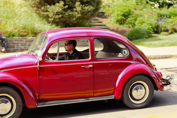 Man with sunglasses is driving a mauve VW bug with whitewall towers with beautiful landscaping in the background