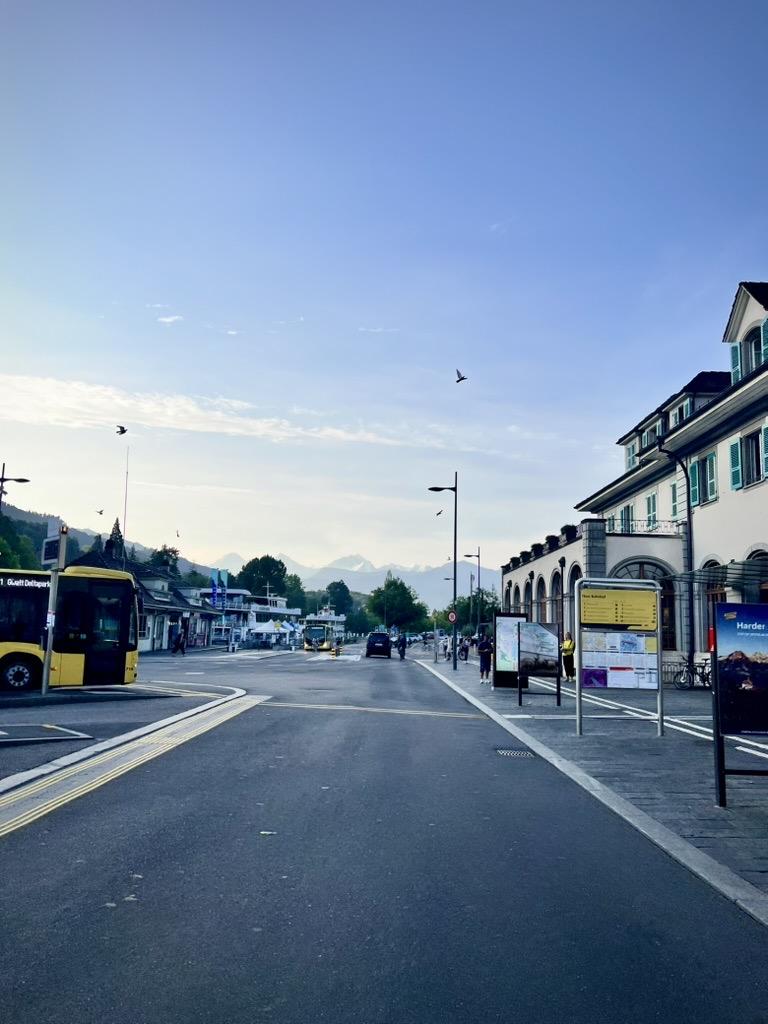 Thun Train Station, Switzerland

A wide, quiet street curves past a white building with green shutters on the right and a bus terminal with a yellow bus on the left, with distant mountains visible on the horizon under a clear morning sky.