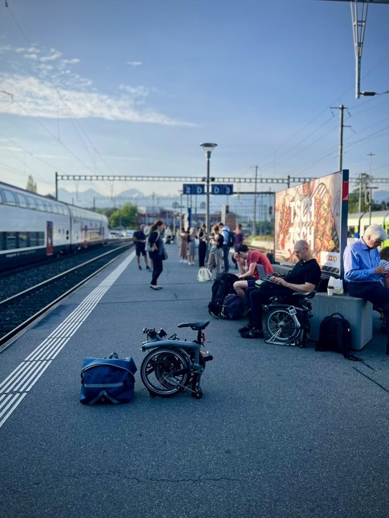 Thun Train Station, Switzerland 

My Brompton and this bag sit in the foreground of a busy train station platform, where commuters wait next to a double-decker train under a blue sky.