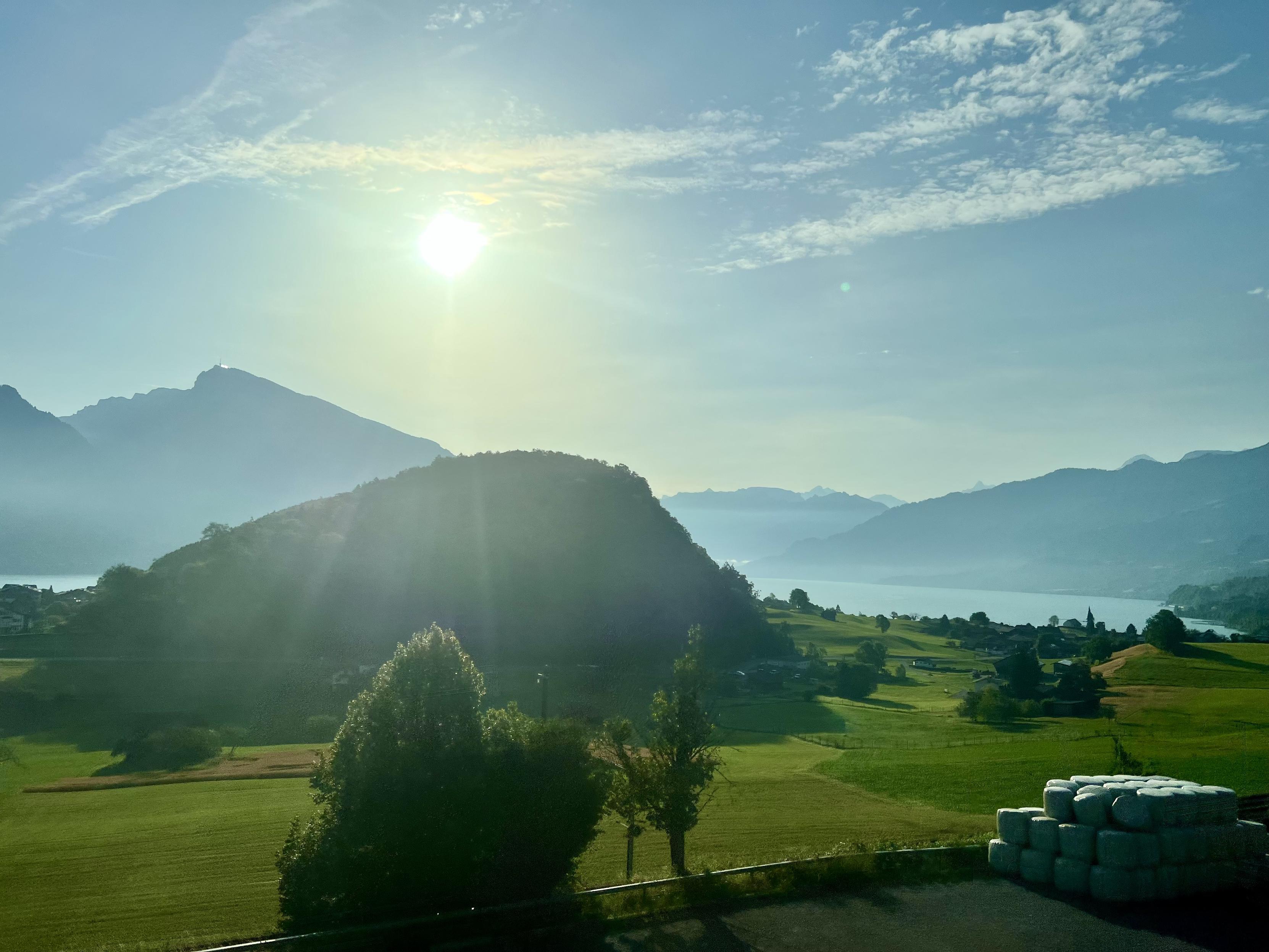 ‎⁨Spiez⁩, ⁨Canton of Bern⁩, ⁨Switzerland⁩
Same location from one of my yesterday photo.

Bright morning sunlight streams over a prominent green hill and surrounding meadows, with silhouetted mountains and a distant lake visible through the hazy, sun-drenched landscape.