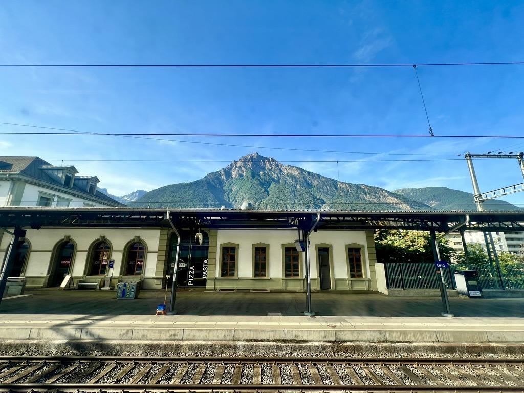 ‎⁨Brigue⁩, ⁨Brig⁩, ⁨Canton of Valais⁩, ⁨Switzerland⁩

A view looking across railway tracks toward a white train station building with arched windows, with a massive, pointed mountain rising steeply directly behind it under a clear blue sky.