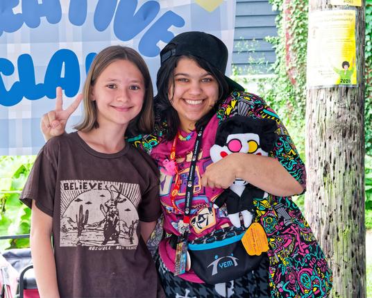 On the right a woman with brightly colored clothes and sideways billed cap and a bag aground her hip and something like a conference lanyard and a plushie in one hand,  she's got her arm behind a girl wearing a brown shirt with (i think) a southwest scene with flying sauces and the word believe in white and she's making a V-sign.  Both are smiling,  I think the sign behind them says something like CREATIVE CLASS and there is a telephone pole behind them with a yellow sign with a green person ho…