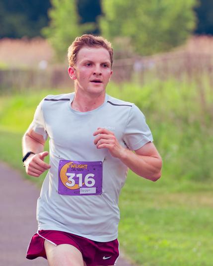 Male runner with short hair has grey shirt and a purple bib with yellow moon with the number 316 with a little corner of pavement on the lower left and green and brown grass and a few trees behind him