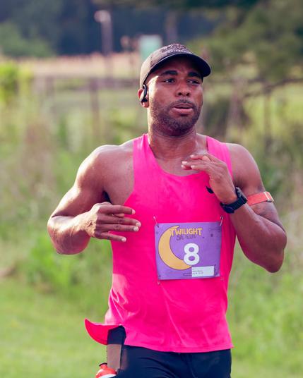 Black man with pinkish-purple tank top, blue shorts and just a hint of a water bottle in his pocket with grass varying from green to brown and short to long in the background