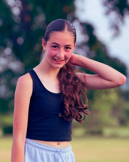 Young woman with dark blue tank top and light blue shorts smiles and looks at me with one hand holding her hair to one side of her neck and an earring in her other ear with a blurry background with light green vegetation below and dark green vegetation above