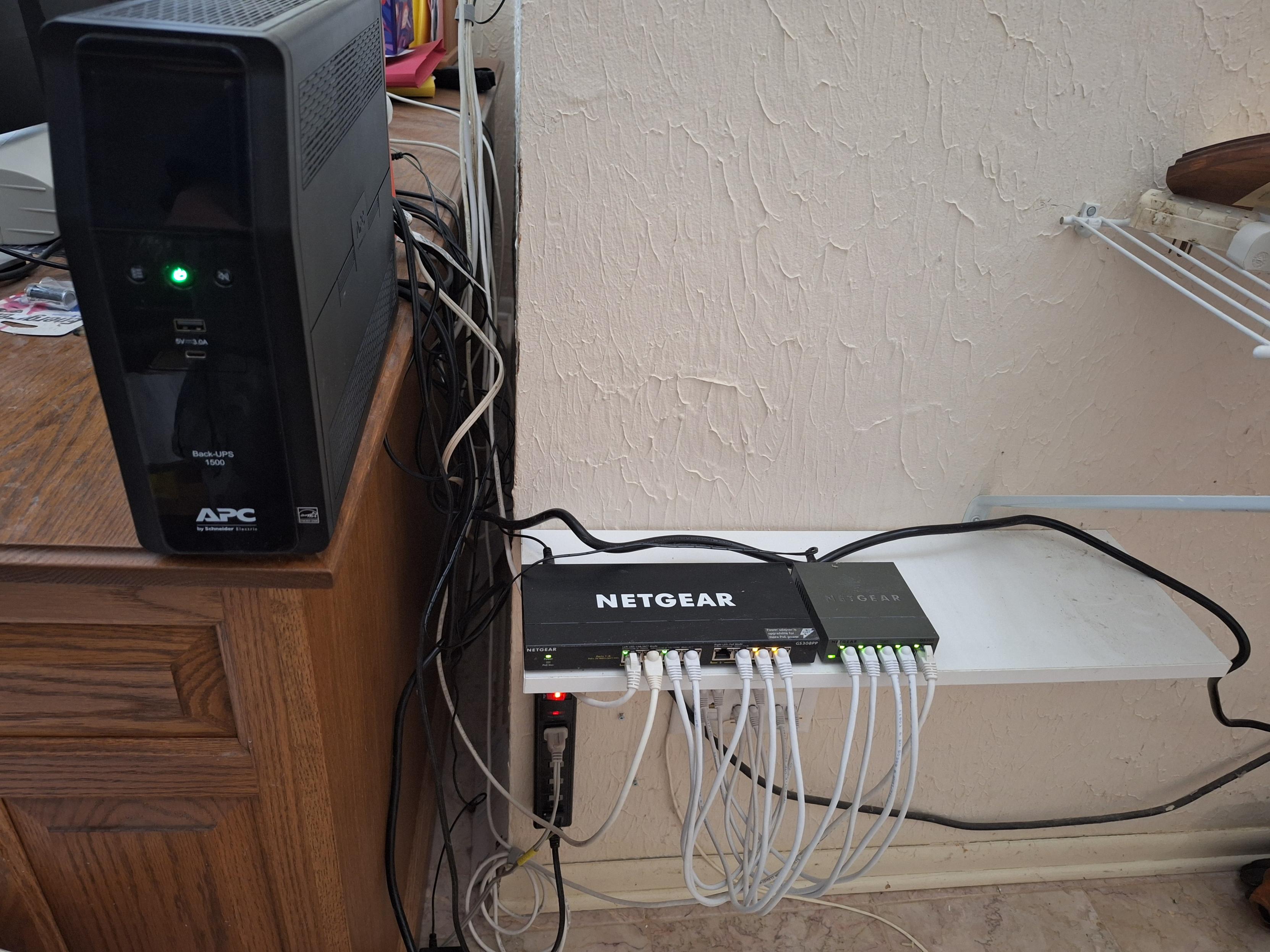 Picture containing ethernet switches.  One is clearly labeled Netgear.  White CAT cables are plugged into their ports.  There is a large black box labeled APC next to the switches.