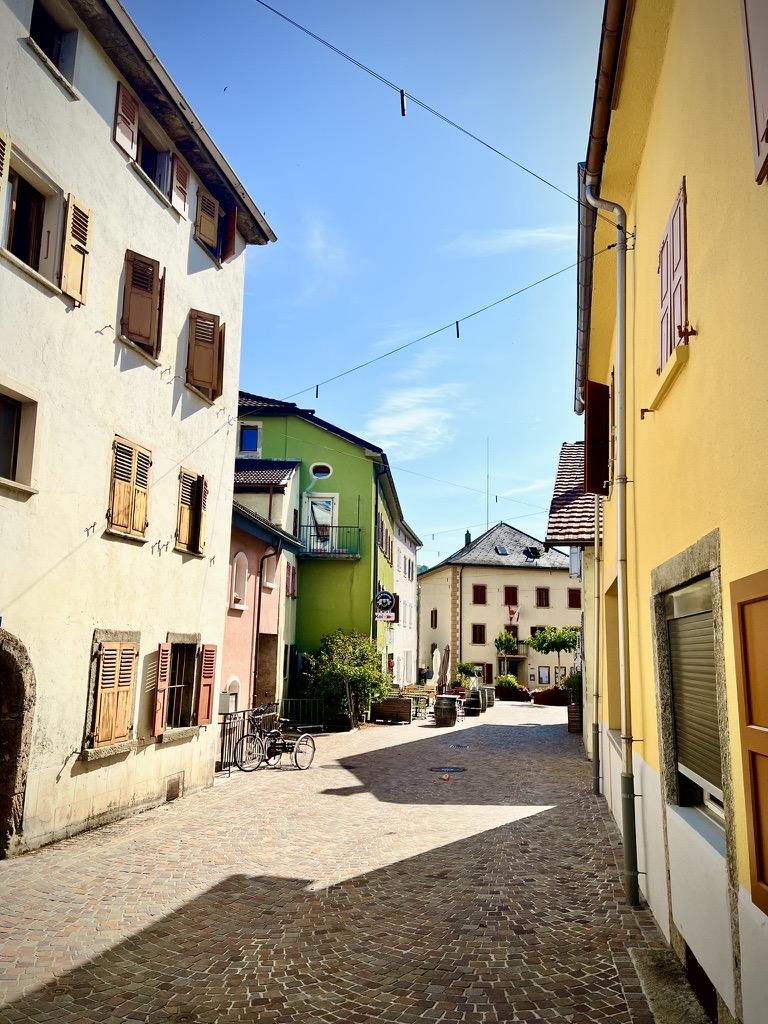 ‎⁨St-Léonard⁩, ⁨Canton of Valais⁩, ⁨Switzerland⁩

A narrow, sunlit cobblestone street curves between multi-story buildings painted in shades of cream, yellow, pink, and vibrant green, many featuring traditional wooden shutters. Deep shadows stretch across the pavement, which leads toward a small open square with outdoor seating in the distance under a clear blue sky.