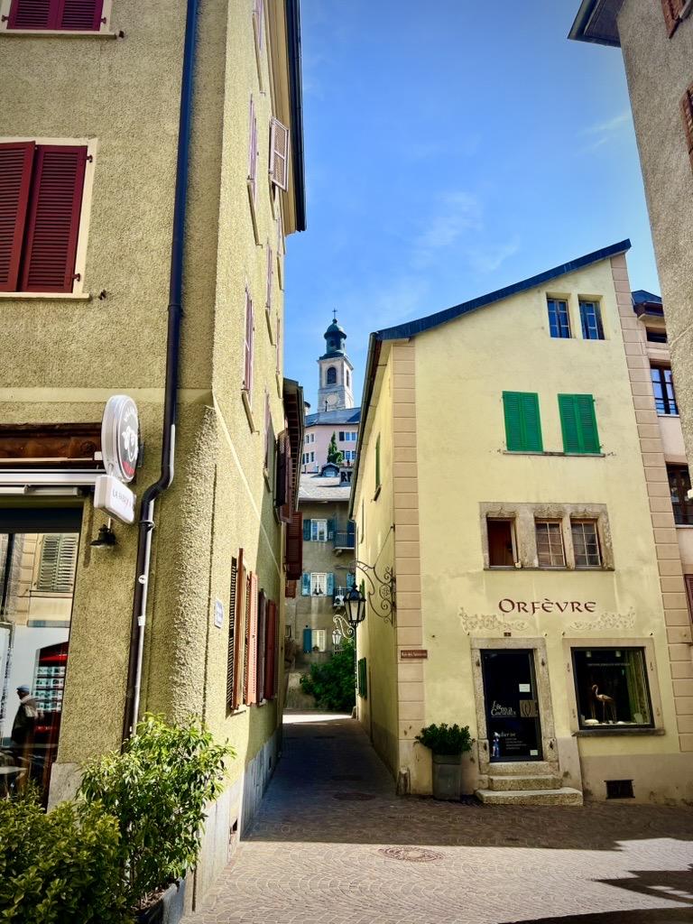 ‎⁨Sion⁩, ⁨Canton of Valais⁩, ⁨Switzerland⁩

A narrow paved alleyway winds between historic yellow and beige buildings with colorful shutters. On the right, a building displays the sign "Orfèvre" above its shop windows. In the background, a historic church tower with a domed roof is framed by the buildings under a bright blue sky.
