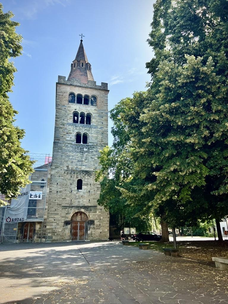 ‎⁨Sion⁩, ⁨Canton of Valais⁩, ⁨Switzerland⁩

A tall, historic Romanesque stone church tower with a pointed spire rises next to large, leafy green trees that cast shadows across a paved courtyard. The tower features tiered arched openings and a wooden door at its base, while a portion of the building to the left is covered in construction scaffolding and protective wrap under a bright, sunny sky.