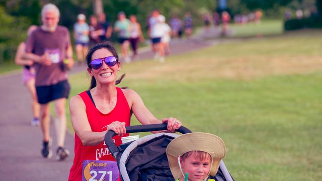 Woman in red tank top and reflective shades pushes a stroller where you can just see the head of a child wearing a wide brimmed cap,  to the right there is a grasy field and to the left and behind her is a stream of blurred out runners