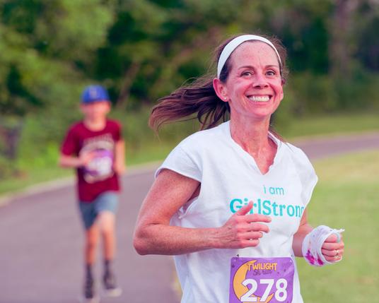 Woman has a a white shirt that reads in blue I am Girlstrong and also a wide headband and something white around one wrist.  As she smiles her hair is flying to the left.  Behind her is a curve trail,  a hedgerow and a blurred out person with the proportions of a child.