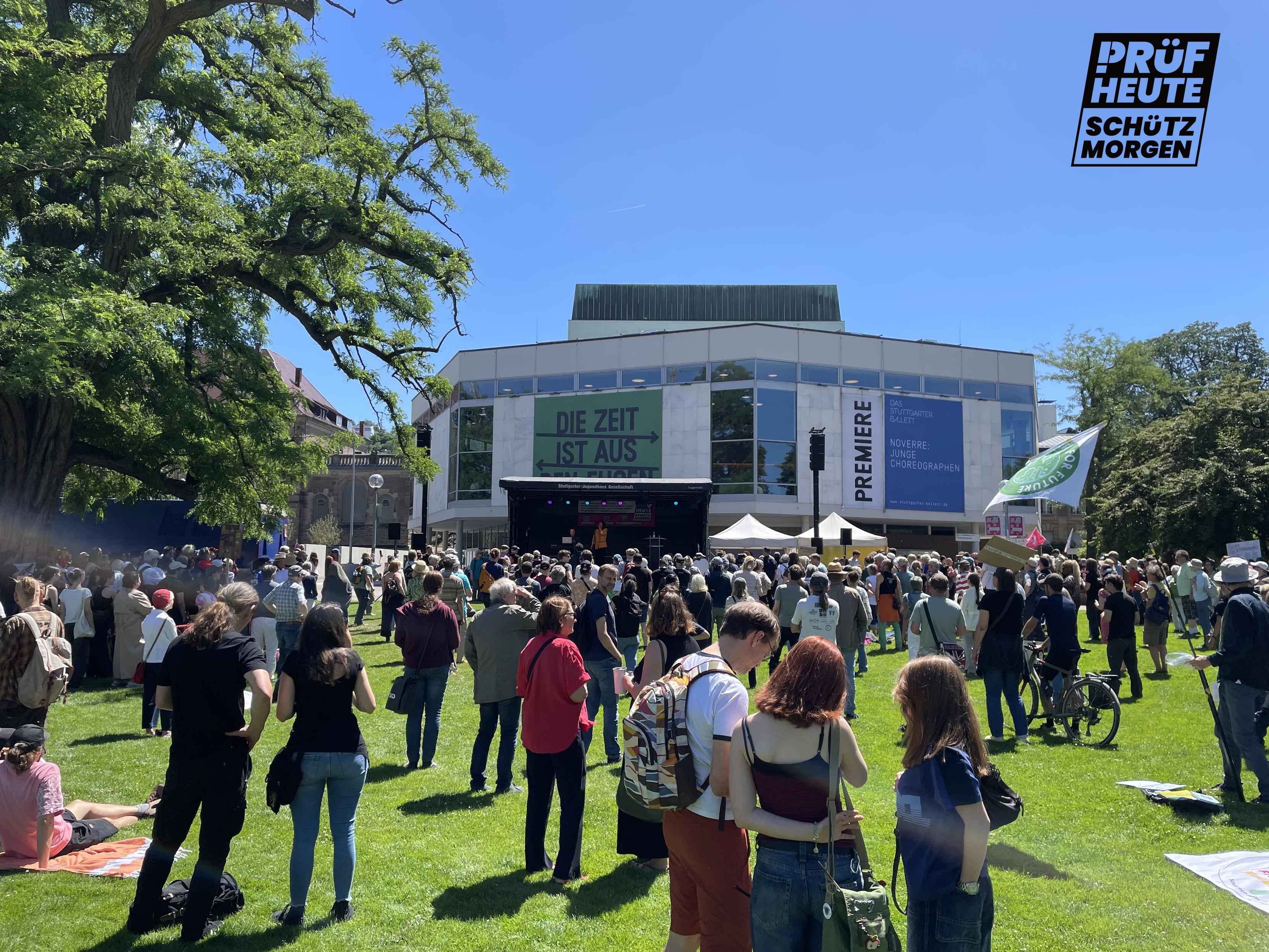 Unter blitzblauem Himmel siehst du das Staatstheater Stuttgart mit den großen Fenstern. Davor haben wir die dunkle Bühne aufgebaut. Links steht einer meiner großen Lieblingsbäume und direkt vor dir sind viele Menschen auf dem grünen Gras. Und wenn du kommst und drei weitere Menschen mitbringst, wird man das Gras nicht mehr sehen...
Unten rechts ist das Logo von PRÜF
"PRÜF
HEUTE
SCHÜTZ
MORGEN"
Das "P" von PRÜF besteht aus einem Ausrufezeichen und einem passenden Bogen am oberen Ende.