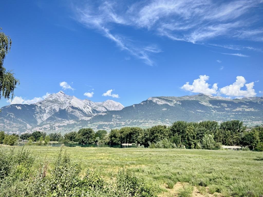‎⁨Domaines des Iles⁩, ⁨Sion⁩, ⁨Canton of Valais⁩, ⁨Switzerland⁩

A wide grassy meadow in the foreground stretches toward a calm lake bordered by a dense line of green trees. In the background, majestic mountain ranges rise beneath a bright blue sky, featuring rugged rocky peaks on the left and forested ridges on the right, all beneath wispy, sweeping clouds.