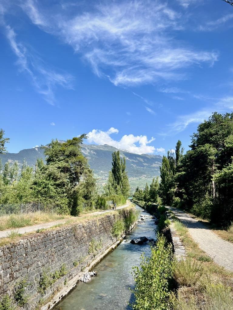 ‎⁨Conthey⁩, ⁨Canton of Valais⁩, ⁨Switzerland⁩

A narrow, stone-lined canal with flowing water is flanked by gravel paths and lush green trees on both sides. In the distance, mountains rise under a vibrant blue sky filled with wispy, sweeping clouds and a few puffy clouds over the peaks.