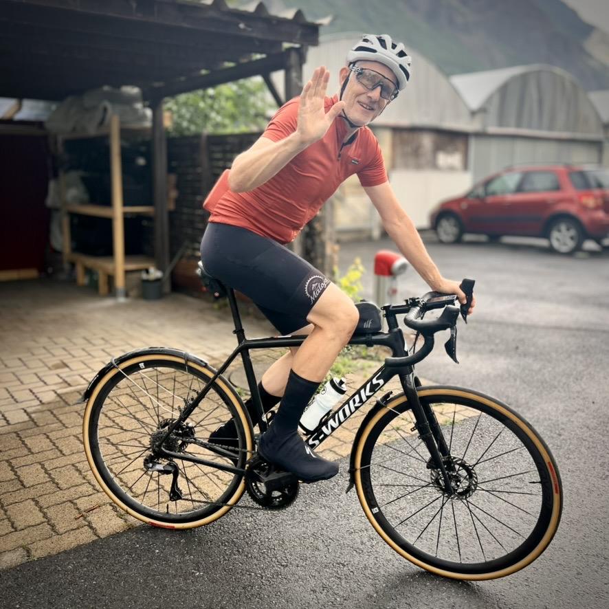 ‎Canton of Valais⁩, ⁨Switzerland⁩

Joakim cycling to work. He wearing a red cycling jersey, black bib shorts, a white helmet, and cycling glasses waves while seated on a black S-Works road bike with tan-wall tires. In the background, a paved area features a wooden carport on the left and a parked red car on the right under soft daylight.