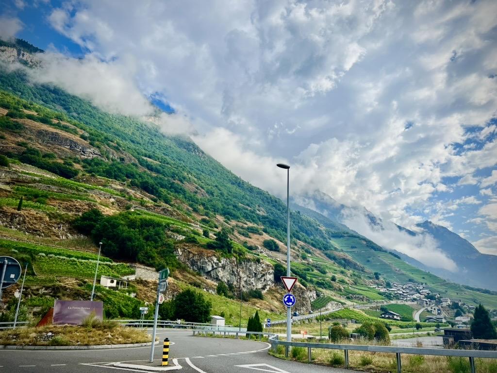 ‎Canton of Valais⁩, ⁨Switzerland⁩

A curved asphalt road and roundabout with traffic signs are framed by a towering, steep hillside on the left, covered in terraced green vineyards, rocky cliffs, and patches of forest. Low-lying clouds cling to the upper mountain slopes, with a small village visible on the lower slopes in the distance under a dramatic sky.