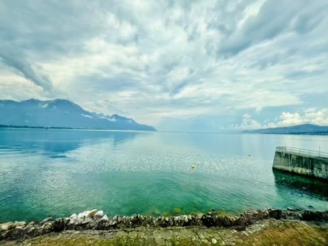 ‎⁨Veytaux⁩, ⁨Canton of Vaud⁩, ⁨Switzerland⁩

An elevated stone embankment and concrete pier overlook a vast, calm lake transitioning from vibrant turquoise near the shore to a deep blue. In the background, rugged mountains stand along the far shoreline beneath a wide, overcast sky filled with textured clouds.