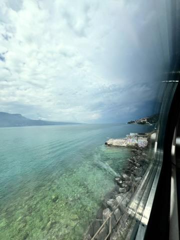 ‎⁨Saint-Saphorin (Lavaux)⁩, ⁨Canton of Vaud⁩, ⁨Switzerland⁩

Seen through a train window showing glass reflections on the right, a rocky shoreline borders a wide lake with clear, turquoise water in the shallows. A small concrete structure covered in colorful graffiti sits on a stone spit, while distant mountains and a hillside town stretch along the horizon under an overcast, cloudy sky.
