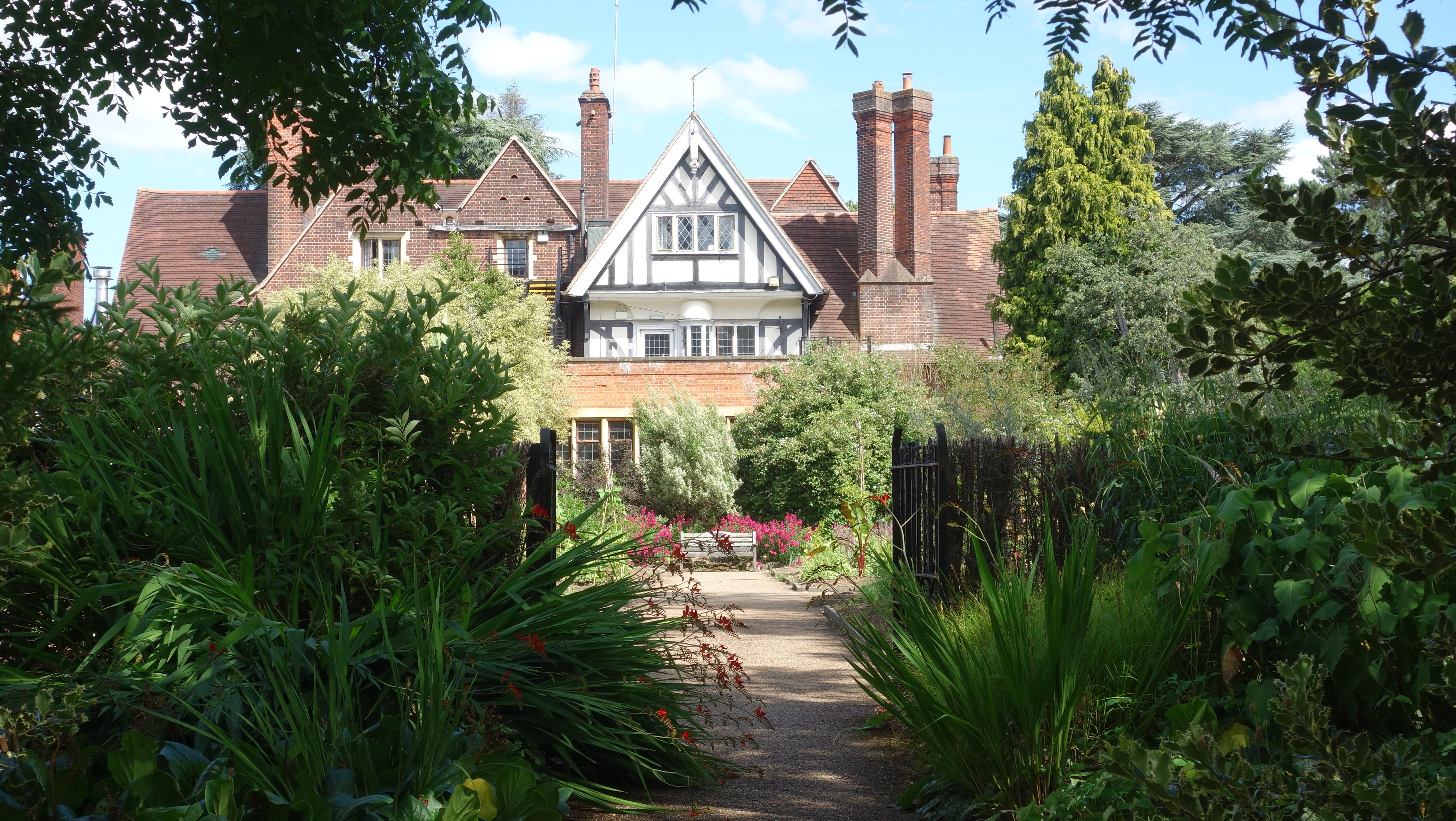 A wide, flat, gravel garden path leads through dark green foliage, overhanging trees, red flowers on strappy leaves and wiry stems (Crocosmia 'Lucifer', perhaps) and a low, open, black, wrought iron gate, into a sunlit garden. Shadows fall across the path, and there are borders with kerbed edges containing lush plants and bamboo wigwams (for sweet peas?).

At the end of the path, there is a simple silver-grey wooden bench with arm rests at either end and horizontal slats at the back. A small metal plaque of some sort is placed centrally on the top slat. The bench is surrounded by crimson flowers, shrubs and trees.

Behind this border is the house; mostly built of red-brick walls and pale mortar, with orange/grey roof tiles and tall orange/grey brick chimneys. At the front and centre of the house, the two upper storeys are black and white and half timbered, with several lead glazed windows, arranged symmetrically above what might be a balcony.

Several ornate chimneys rise well above the main roof line, with clay pots on their tops, some with vents to increase the updraught. The pair at the front right of the house are square sectioned, but rotated 45 degrees (around a vertical axis) with respect to the house. Clear sky can be seen between the two chimneys.

The half-timbered roof line is triangular, rising to a central point, as do several other roof features. More windows can be seen near ground level, and on the first floor, to the left. Behind the house, more large trees are just visible, reaching up into a sky-blue sky, with a few white clouds.

The overall feeling is one of calm and warmth.