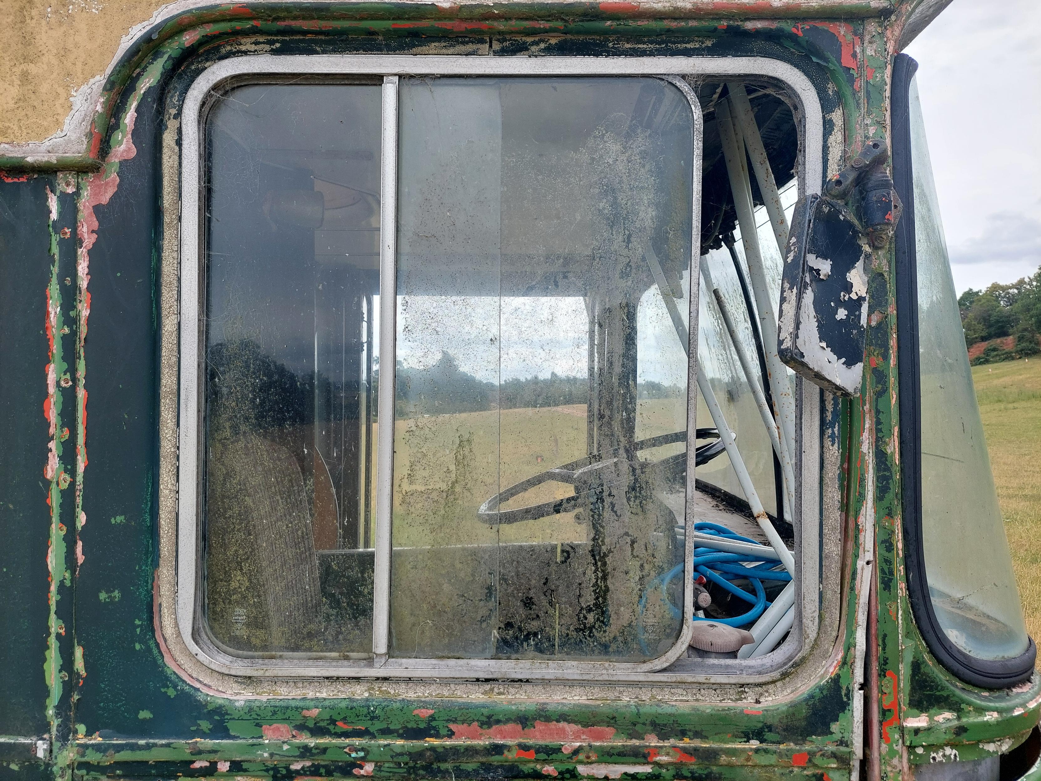 Close-up of the side window of a derelict bus standing in the middle of a field. Flaking layers of green, cream and red paint expose years of weathering, while the dirty, clouded glass offers a glimpse of the stripped interior, with the steering wheel, metal supports and a coil of bright blue hose still inside. Beyond the window, rolling pasture and distant trees are faintly reflected, contrasting with the bus's slow decay.