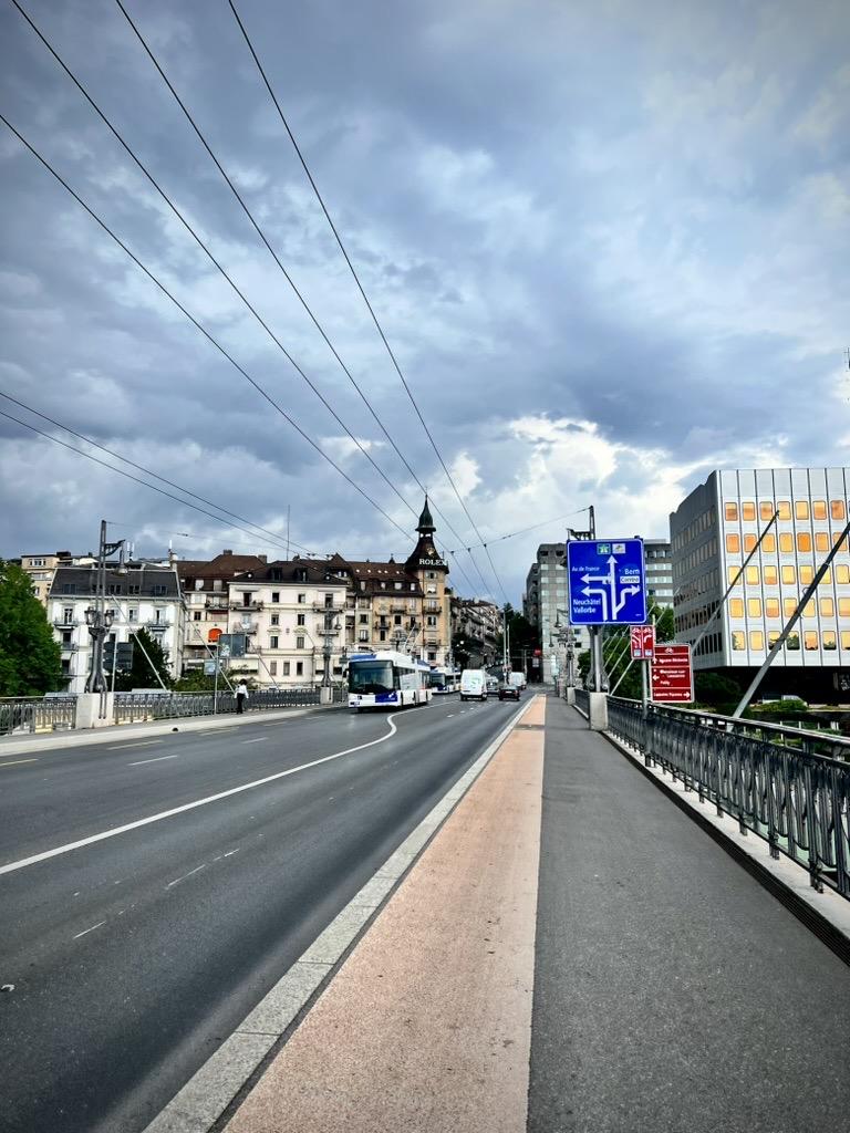 ‎⁨Lausanne⁩, ⁨Canton of Vaud⁩, ⁨Switzerland⁩

A wide road bridge featuring a pedestrian walkway on the right and overhead trolleybus cables stretching across. Vehicles, including a public bus, travel along the roadway, which is lined with blue and red directional signs. In the background, a historic clock tower and various buildings stand beneath a dramatic, heavily clouded sky.